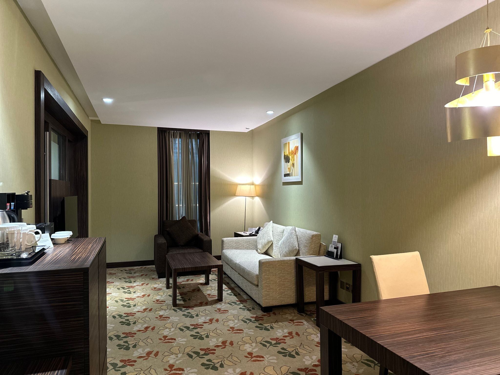 A hotel room with a sofa, armchair, small table, and a desk with a lamp.