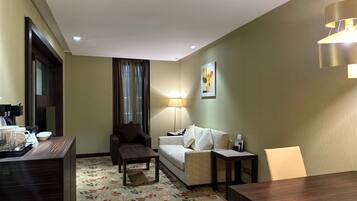 A hotel room with a sofa, armchair, small table, and a desk with a lamp.
