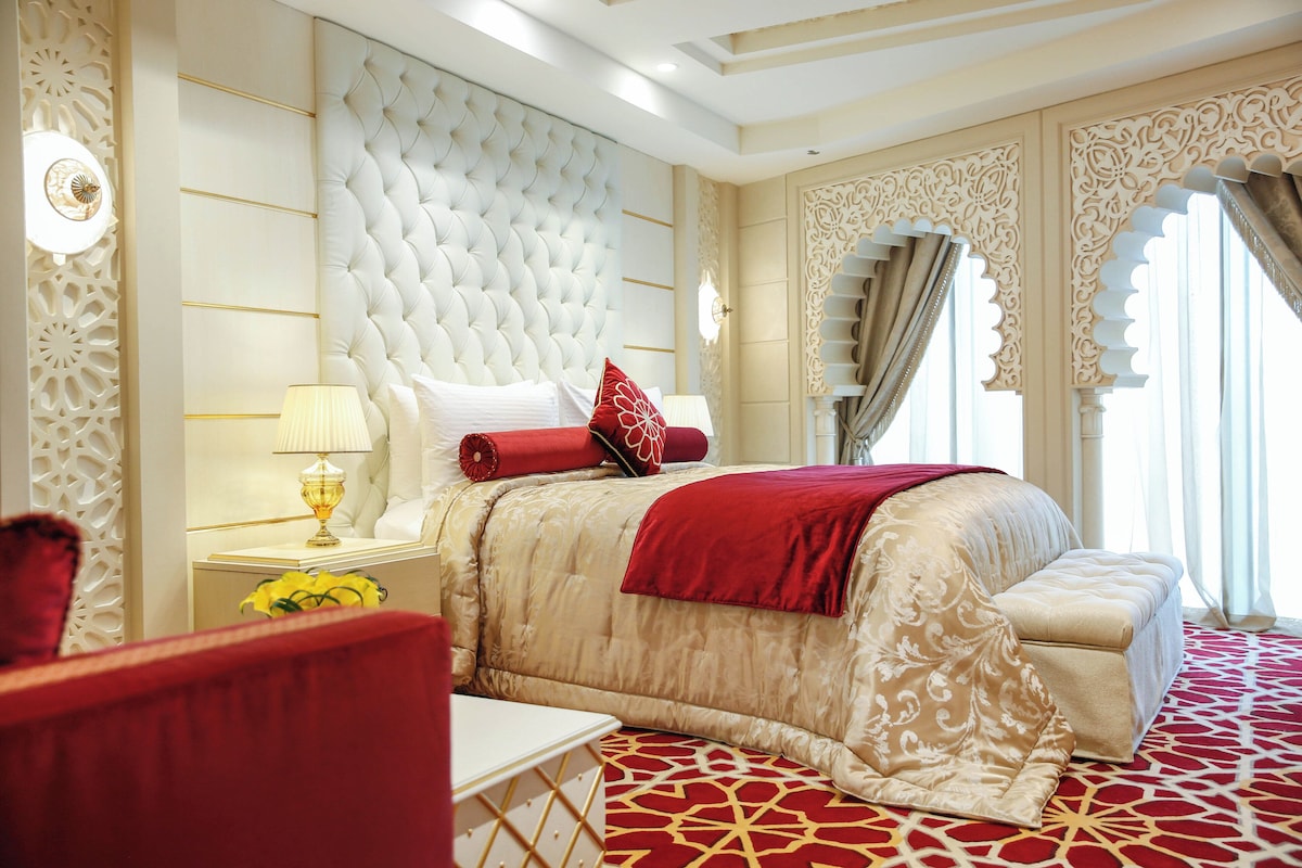 A luxurious bedroom with a tufted headboard, a red throw blanket, and a patterned carpet.