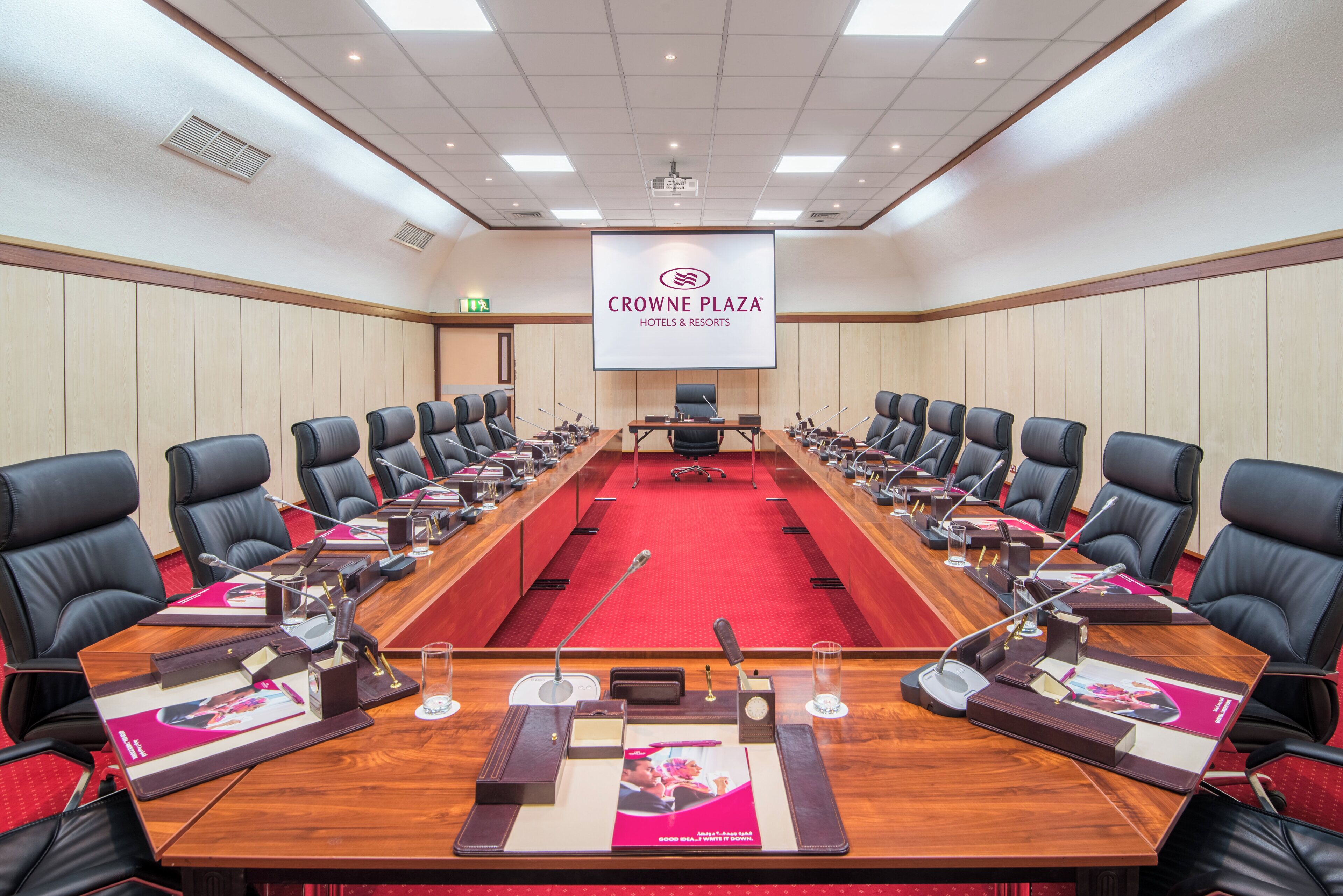 A conference room with a long table, multiple chairs, microphones, and a screen displaying the Crowne Plaza Hotels & Resorts logo.