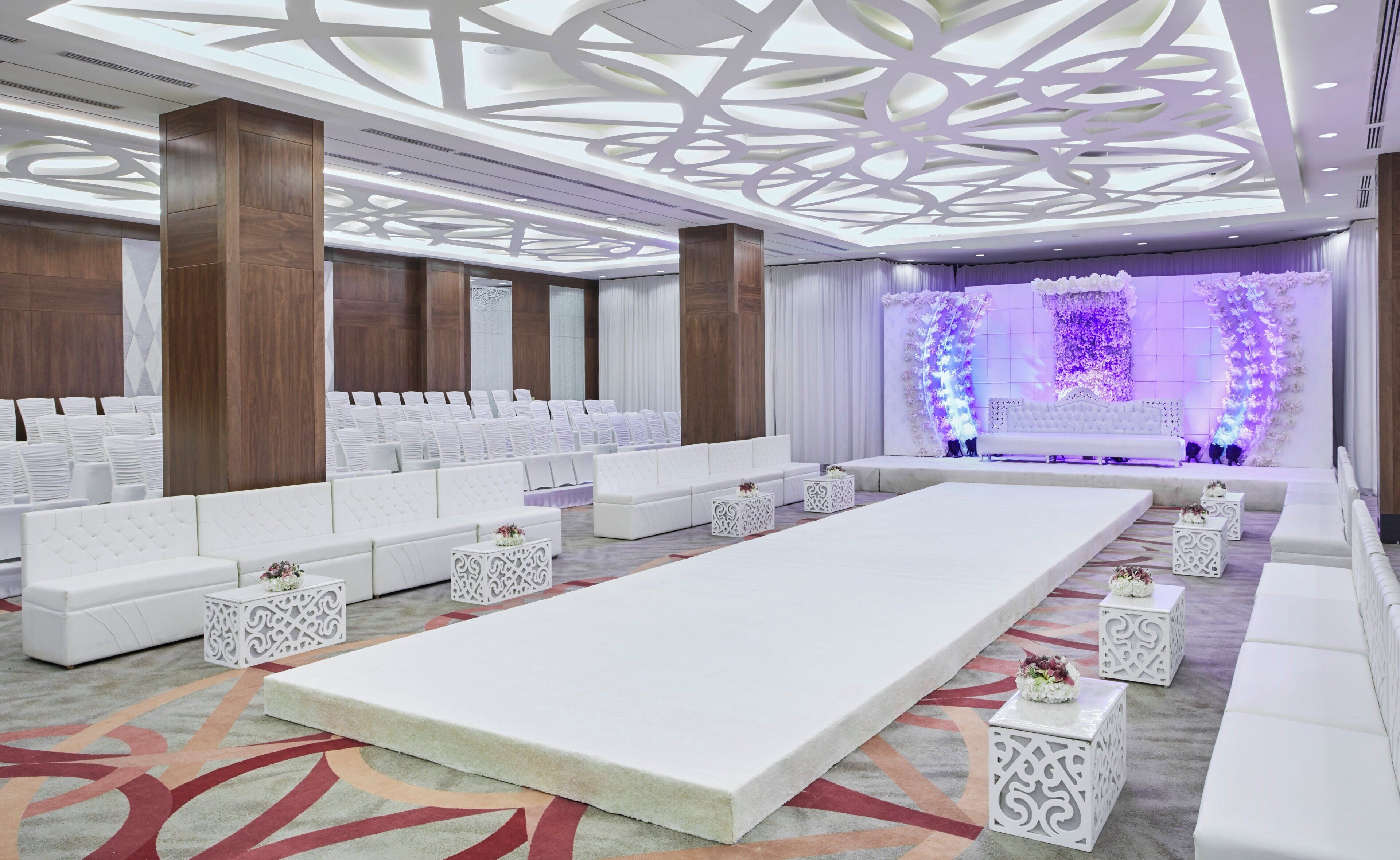 A spacious room with a long white sofa, white chairs, and a stage with floral decorations.