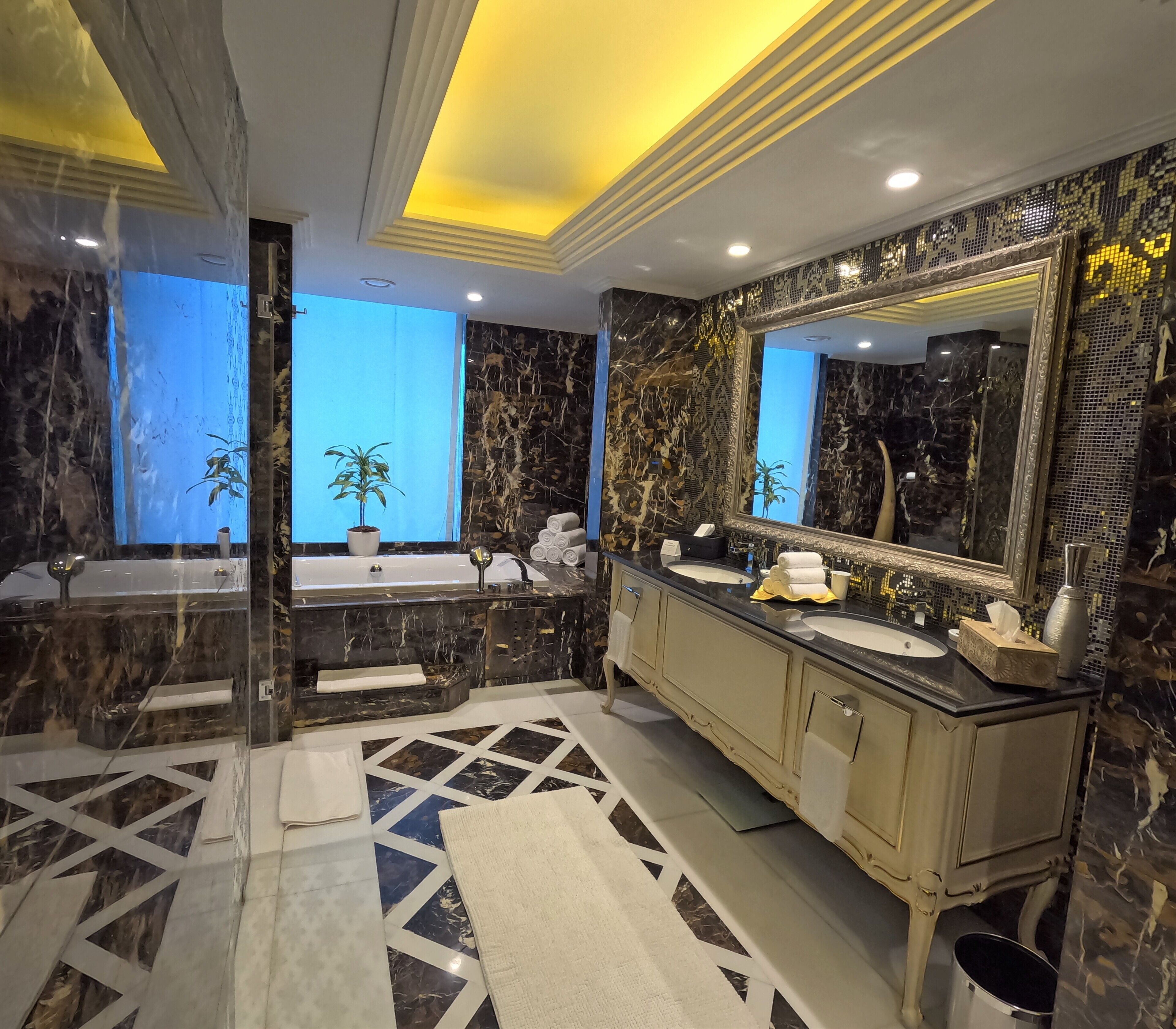 Une salle de bain luxueuse dotée d’un double lavabo, de plans de travail en marbre, d’un grand miroir et d’un sol au motif géométrique.