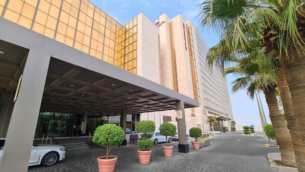 A modern hotel with a glass facade, a covered entrance, and potted trees in front. - Crowne Plaza Kuwait Al Thuraya City by IHG (Farwaniya)