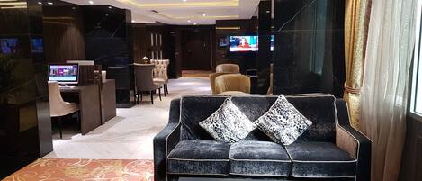 A modern hotel lobby with a dark sofa, a glass coffee table, and a dining area with a TV.