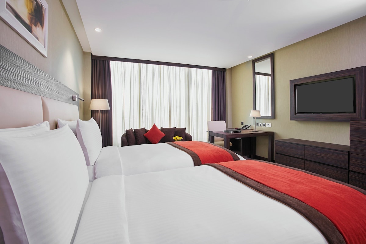 A hotel room with two beds, a desk, a TV, and a window with curtains.