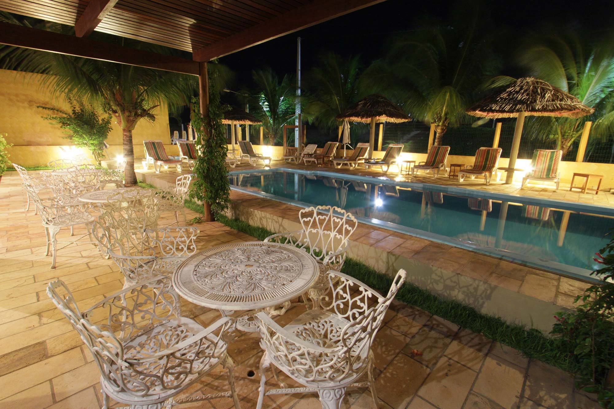 An outdoor pool area with lounge chairs and tables, surrounded by palm trees and a covered seating area.