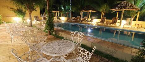 An outdoor pool area with lounge chairs and tables, surrounded by palm trees and a covered seating area.