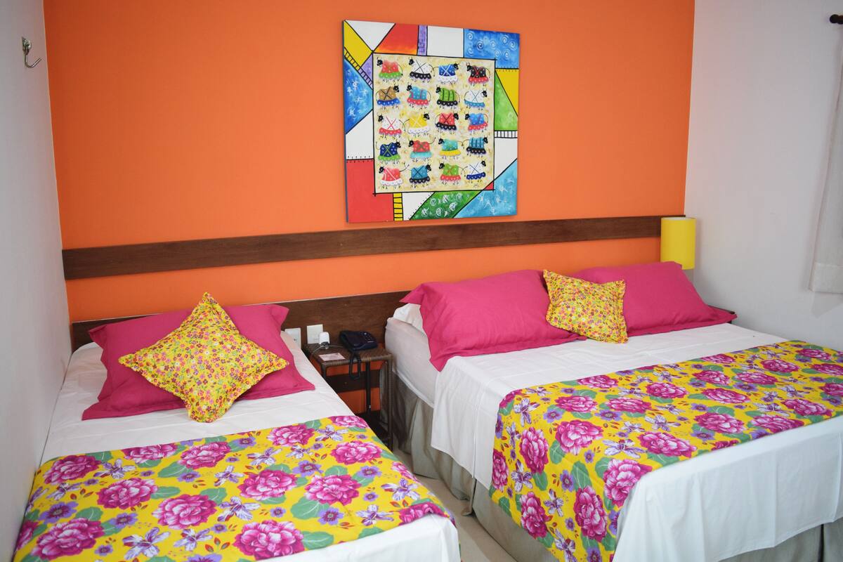 Habitación de hotel con dos camas, cada una con colchas estampadas y almohadas rosas. En la pared naranja sobre las camas, hay una obra de arte colorida.