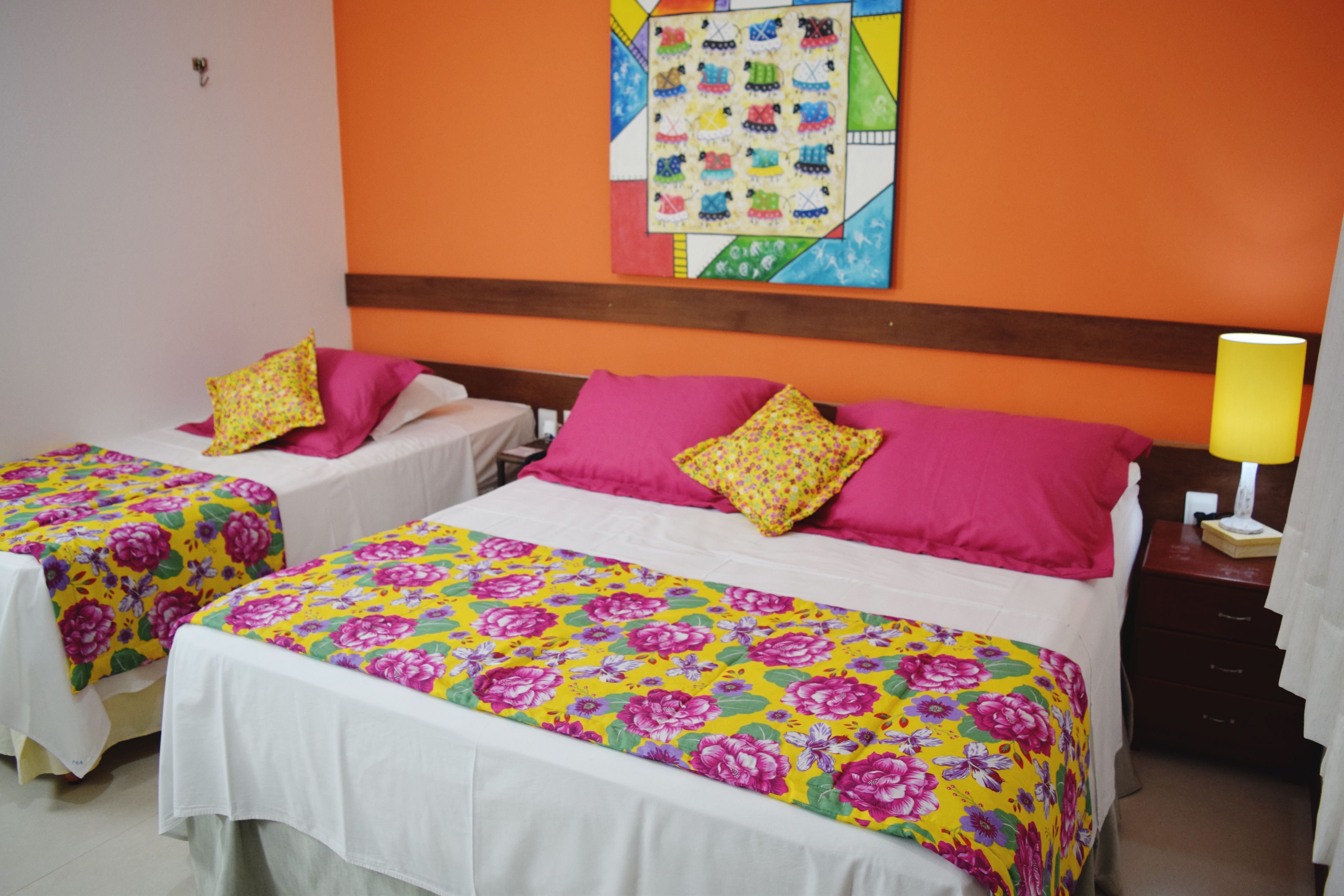 A hotel room with two single beds, each with a floral bedspread and pink pillows. There is a colorful artwork on the wall above the beds and a nightstand with a lamp to the right.