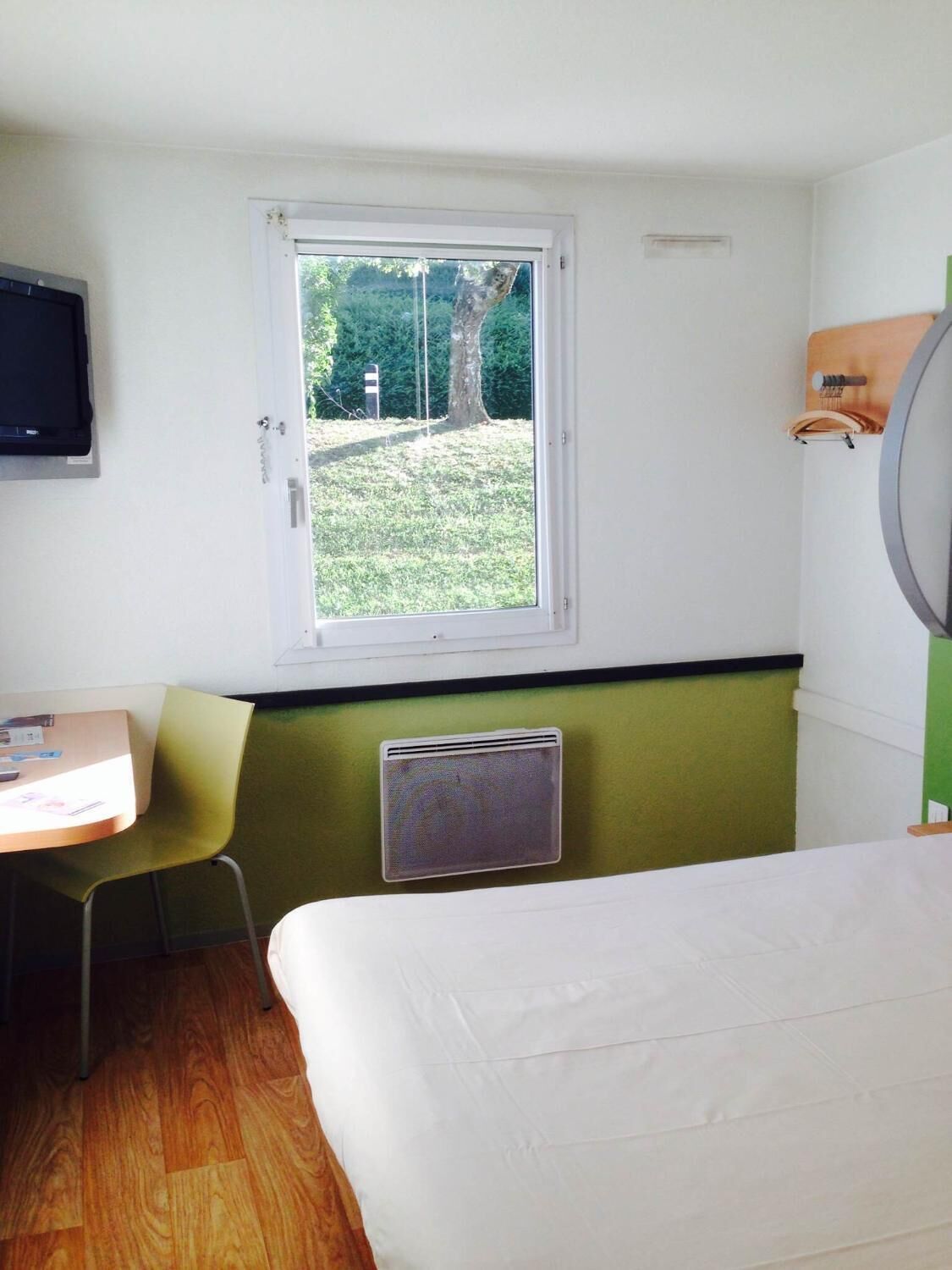 Ein Zimmer mit Bett, Schreibtisch, Stuhl, einem Fenster mit Blick auf Grünflächen und einem Wand-Fernseher.