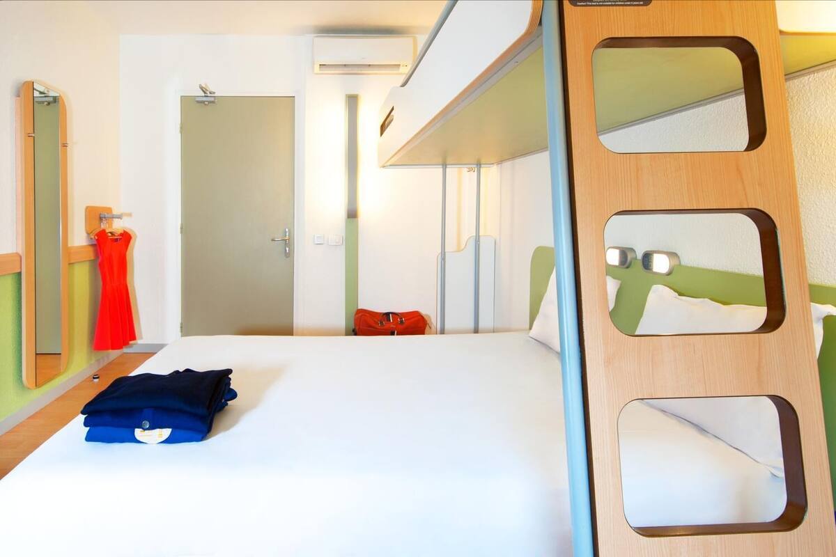 Ein Hotelzimmer mit Etagenbett, einem Spiegel mit Ausschnitten, einem Bett mit weißer Bettdecke, einem roten Kleid, das an der Wand hängt, und einer roten Tasche auf dem Boden.