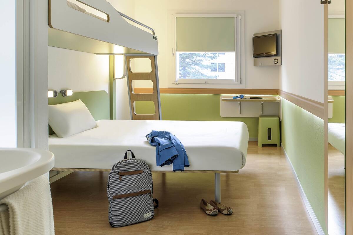 Ein kleines Zimmer mit Etagenbetten, Schreibtisch, Stuhl, kleinem Tisch und Fenster.