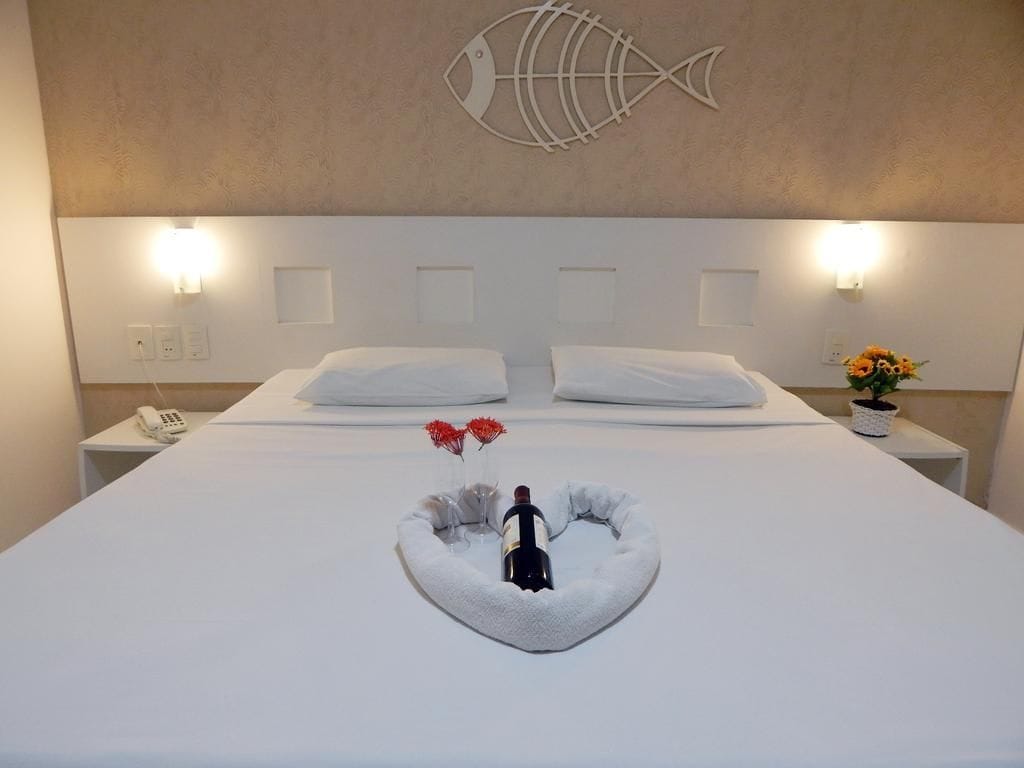 Quarto de hotel com uma cama grande, cabeceira branca e uma arte de parede decorativa com peixes.