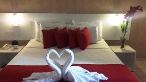 A hotel room with a bed, red pillows, a heart-shaped towel arrangement, a vase with flowers, and a nightstand with a phone and a small plant. - Perolas Do Mar Pousada (Maceio)