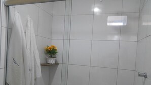 A bathroom with white tiled walls, a glass shower door, and a shelf with a vase of yellow flowers. - Perolas Do Mar Pousada (Maceio)