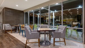 A modern indoor space with a dining area featuring grey upholstered chairs and a wooden table, large windows offering views of the interior, and a tiled floor. - Home2 Suites by Hilton Orlando/International Drive South (Orlando)