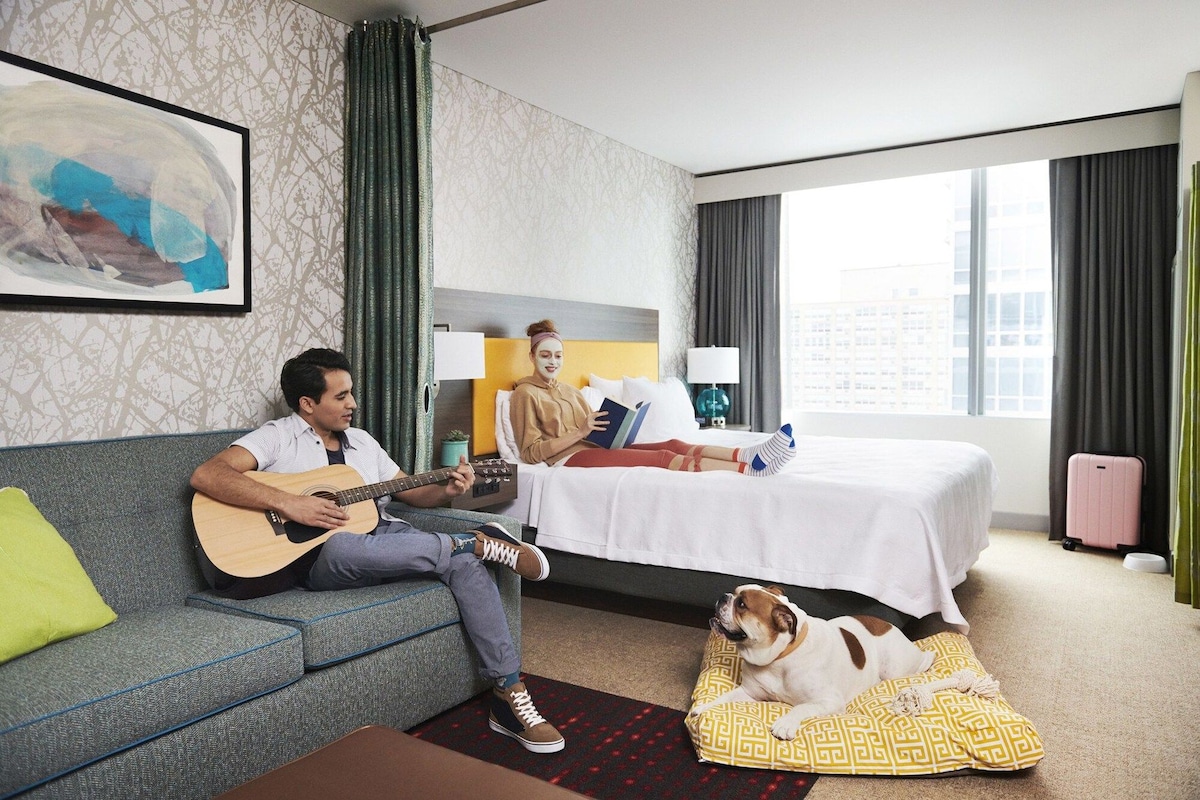 Una habitación de hotel con una cama, un sofá, un perro y una persona tocando la guitarra.