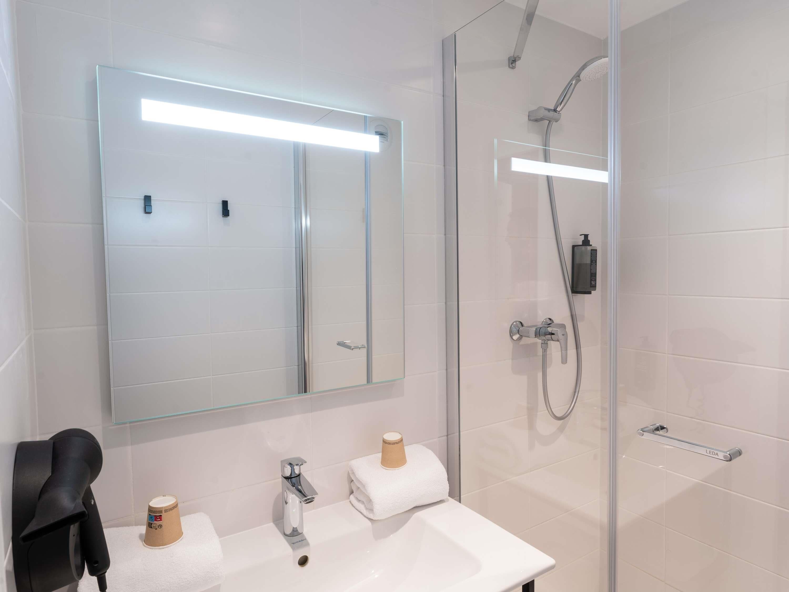 Un baño moderno con cabina de ducha de vidrio, un espejo grande y un lavabo blanco con una toalla y una taza.