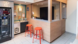 A reception area with a wooden counter, two red stools, a Saeco coffee machine, and a 'Heart of Coffee' sign. - ibis budget Rennes Cesson (Cesson-Sevigne)