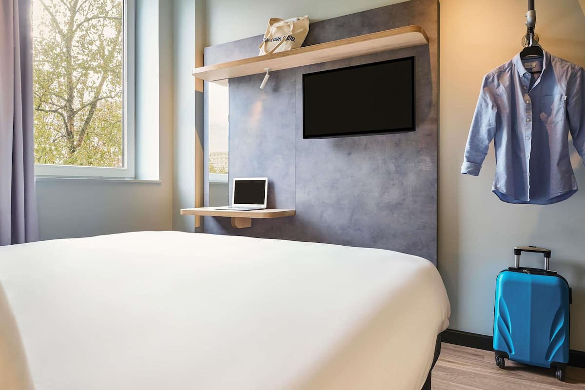 A hotel room with a bed, a desk with a laptop, a TV mounted on the wall, a hanging coat, and a suitcase.