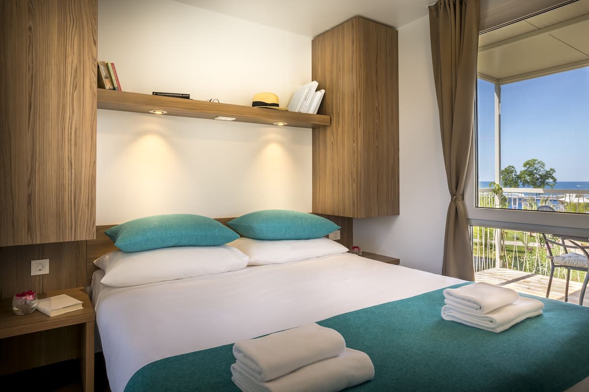 Un lit bien fait, avec une literie blanche et turquoise, une tête de lit en bois et une vue sur la mer par une grande fenêtre.