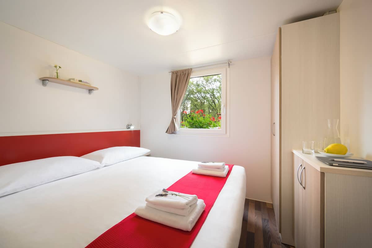 Un lit bien fait, avec du linge de lit blanc et des touches de rouge, une petite étagère ornée d’objets décoratifs et une fenêtre donnant sur un paysage verdoyant.