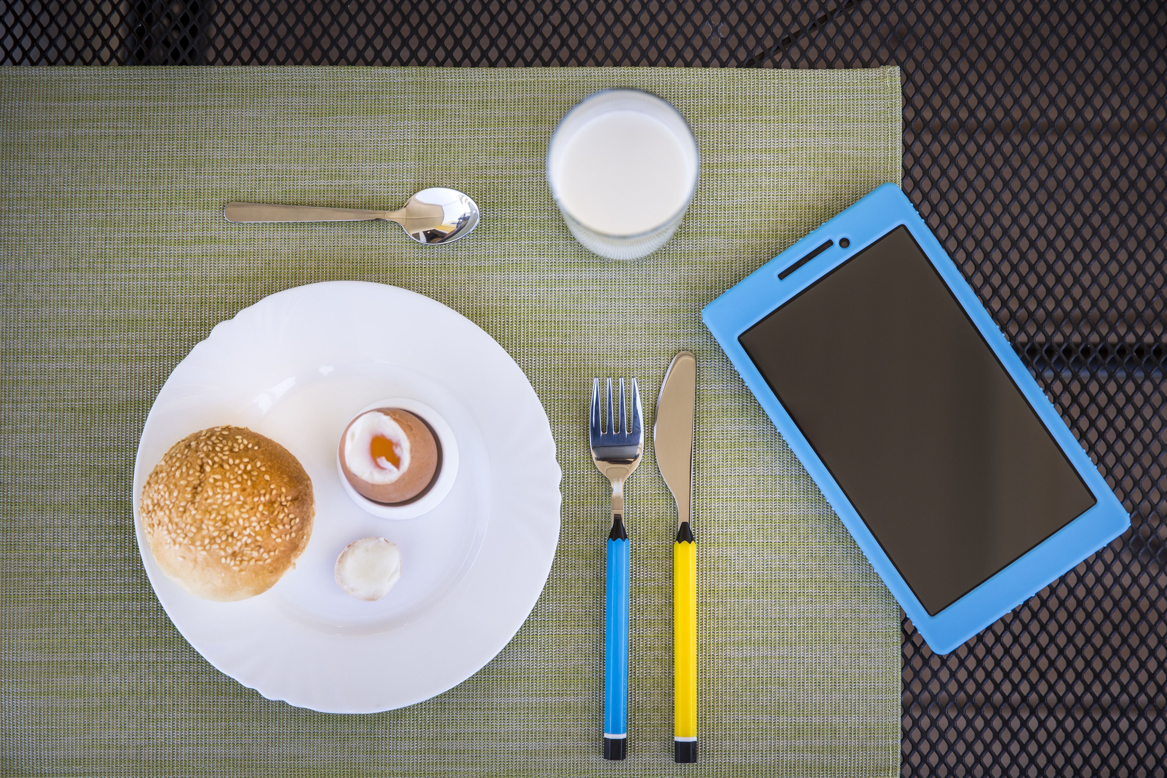 A meal with a bun, a small dessert, and a glass of milk on a table with a tablet, fork, and knife.