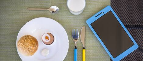 Plato con un panecillo, un postre pequeño y un vaso de leche sobre una mesa con una tableta, un tenedor y un cuchillo.