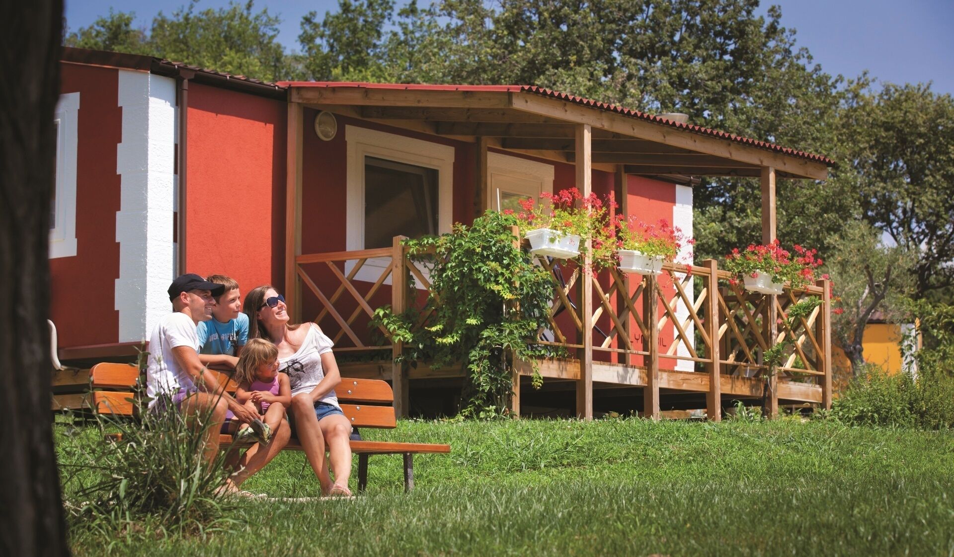 A wooden bench with people sitting on it in front of a red and white building with a wooden porch and potted plants.