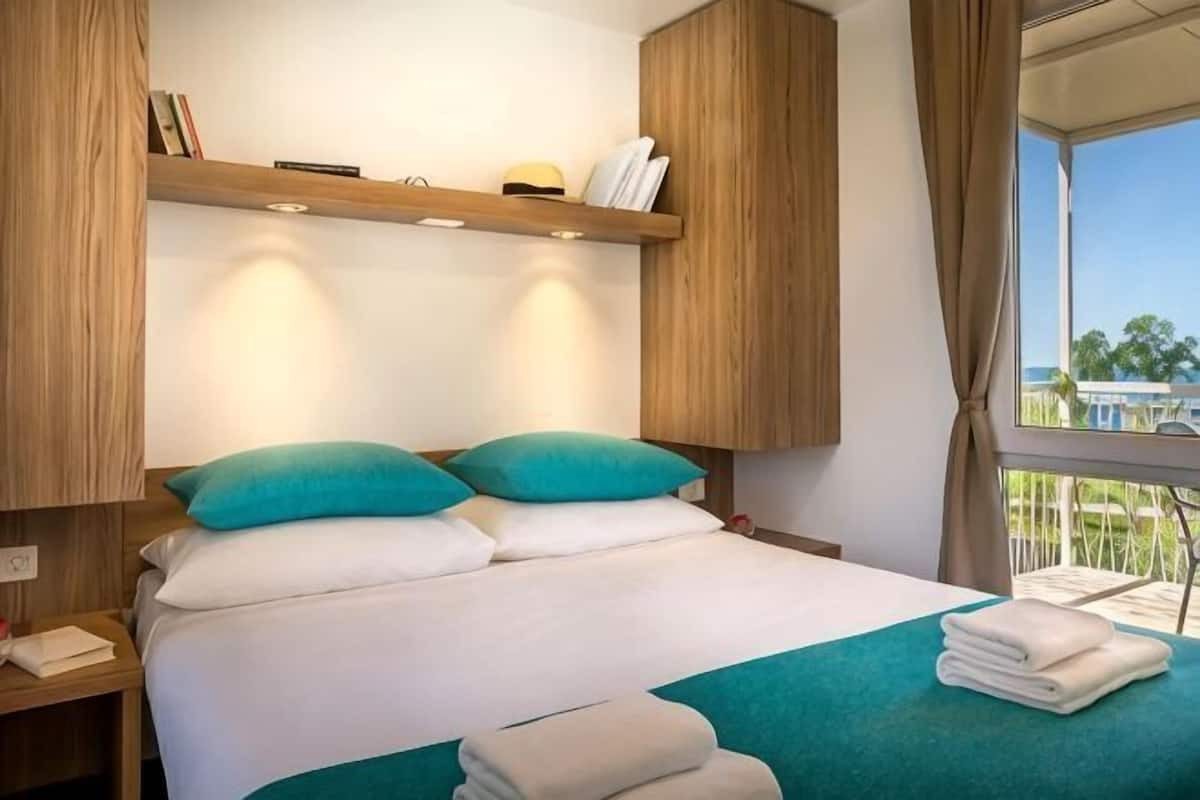 Une chambre d’hôtel avec un lit, des oreillers, une table de chevet, une fenêtre donnant sur un paysage agréable et une étagère avec divers objets.