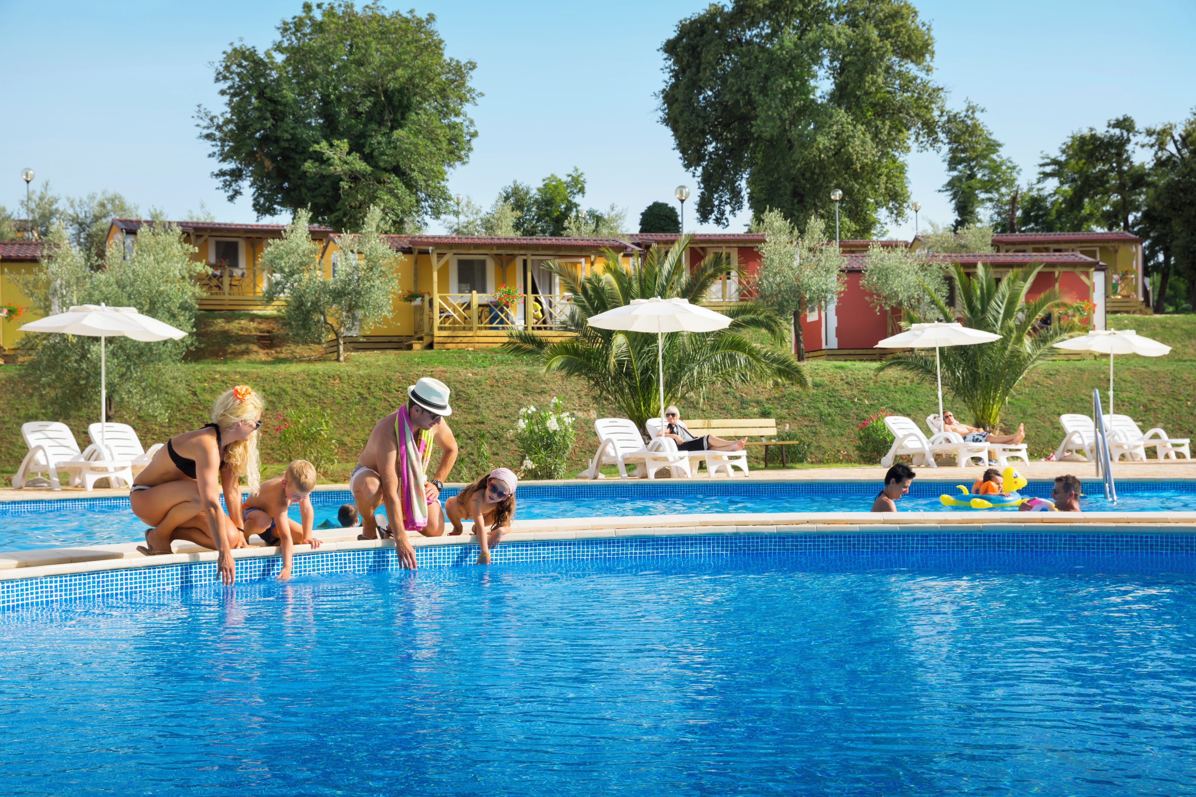A swimming pool area with lounge chairs and umbrellas, surrounded by yellow buildings and greenery.