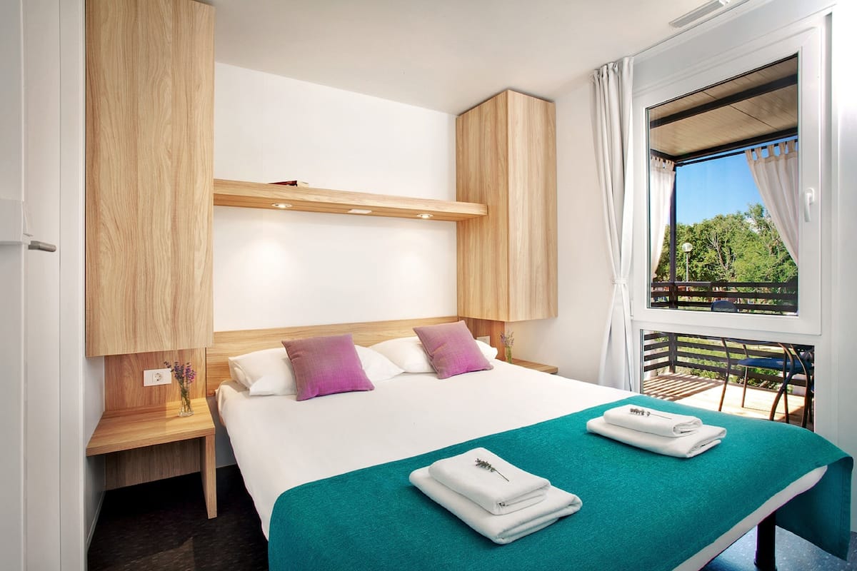 Une chambre d’hôtel moderne avec un grand lit, une tête de lit en bois, une fenêtre avec des rideaux et un balcon offrant une vue agréable.