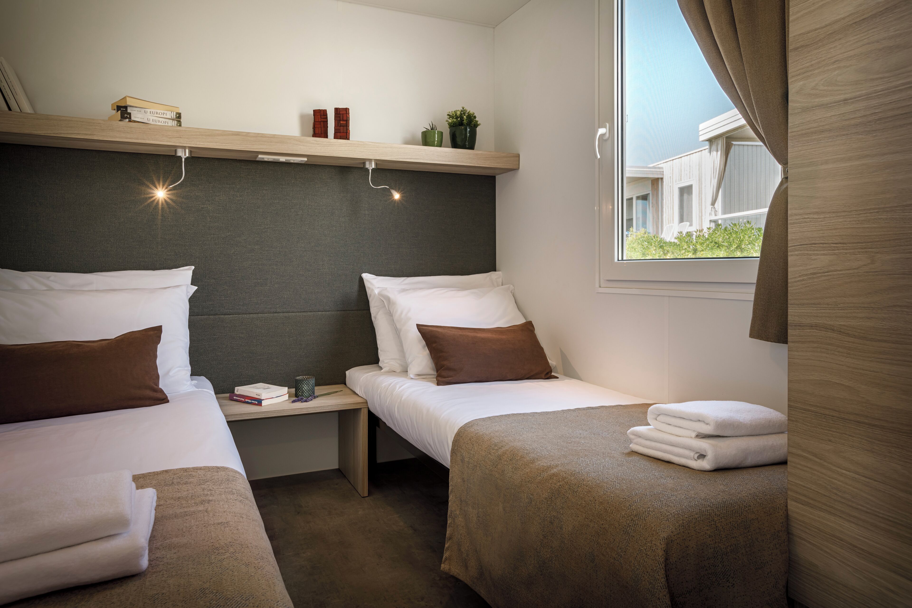 A hotel room with two beds, a bedside table, a shelf with decorative items, and a window with curtains.