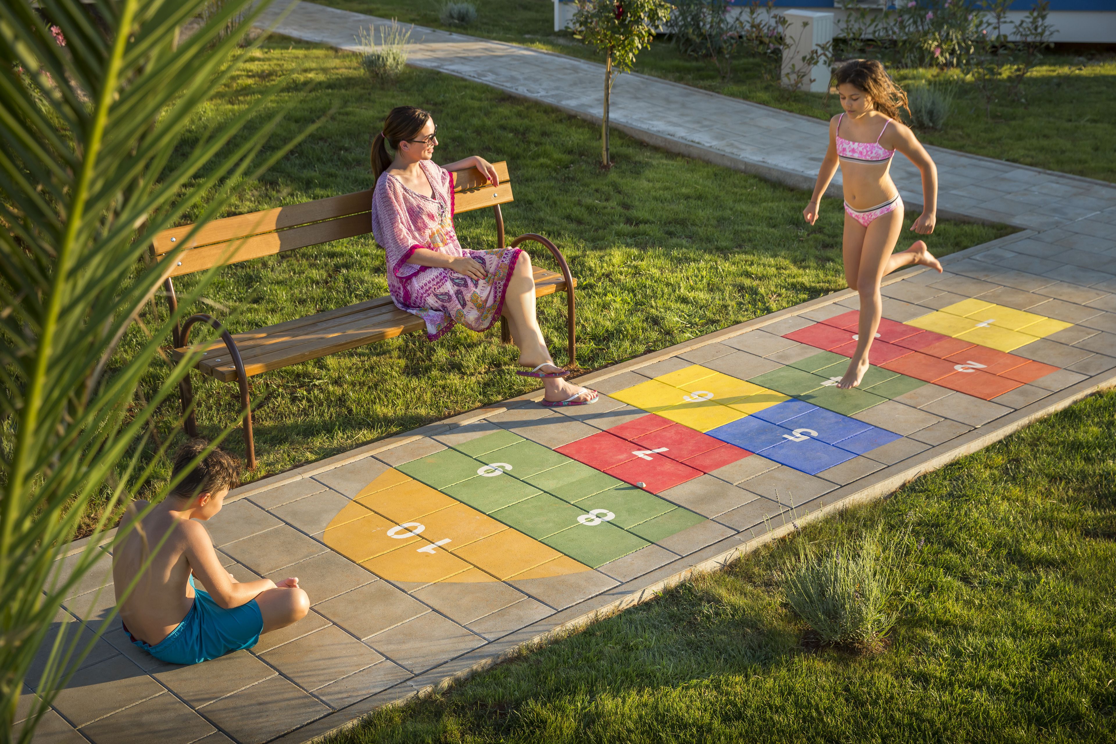 Un parc avec un banc en bois, un jeu de marelle et un enfant qui joue.