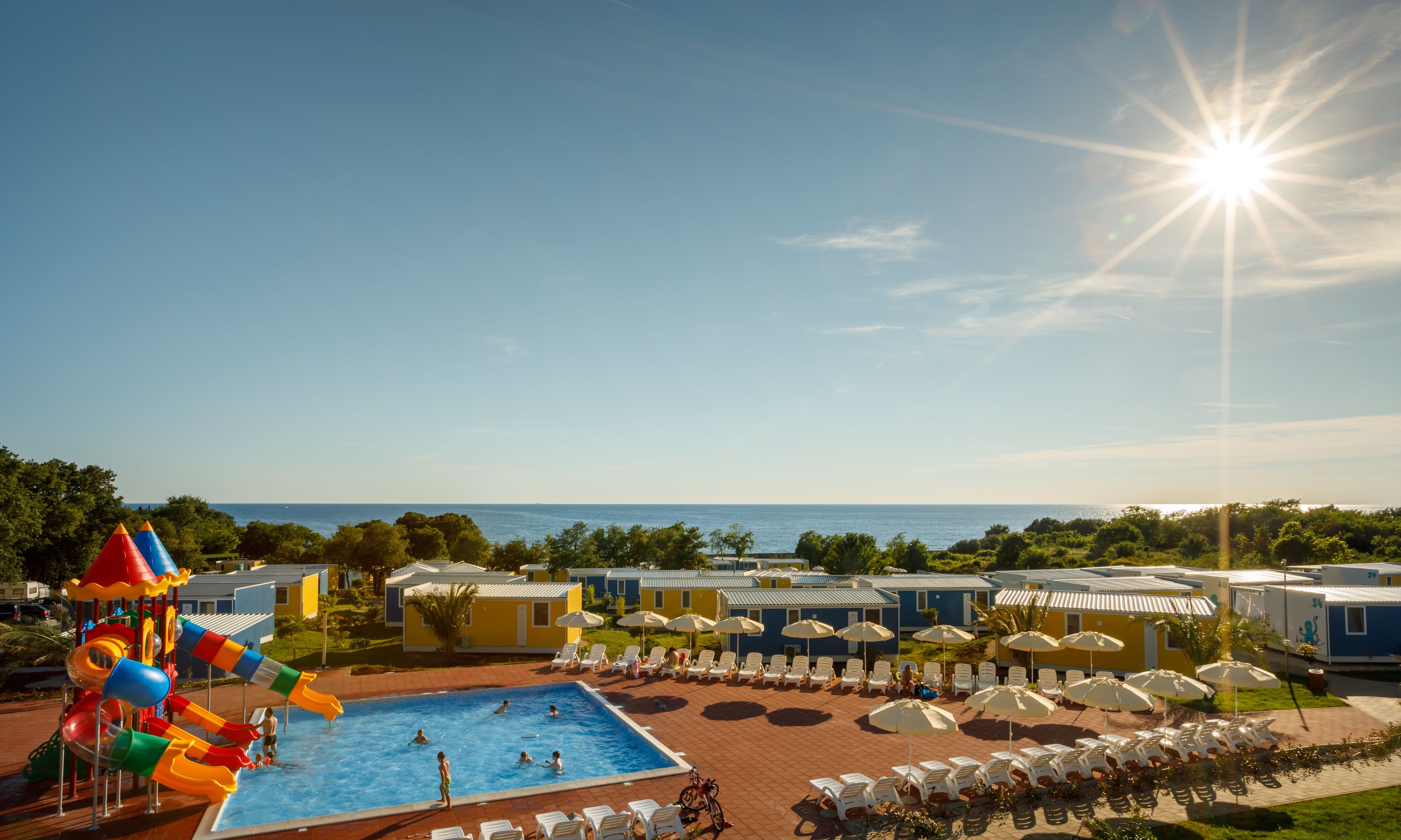 A swimming pool with water slides, sun loungers, and umbrellas, surrounded by beach houses.