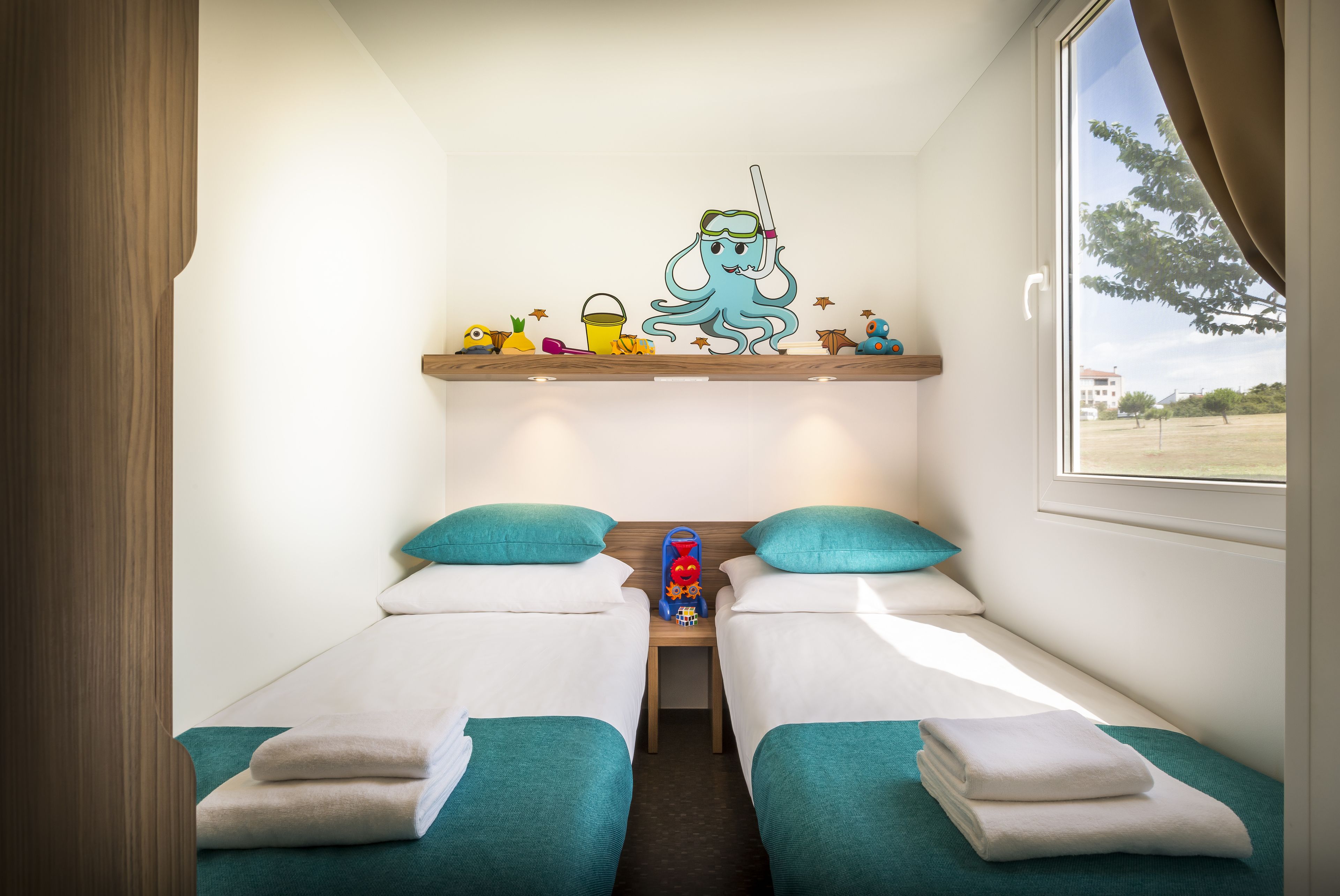 Une chambre d’hôtel avec deux lits, une literie de couleur turquoise, une petite table avec des jouets et une fenêtre donnant sur l’extérieur.