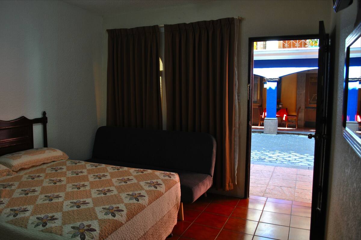 A hotel room with a bed, a sofa, and a view of the outdoor area through a door.