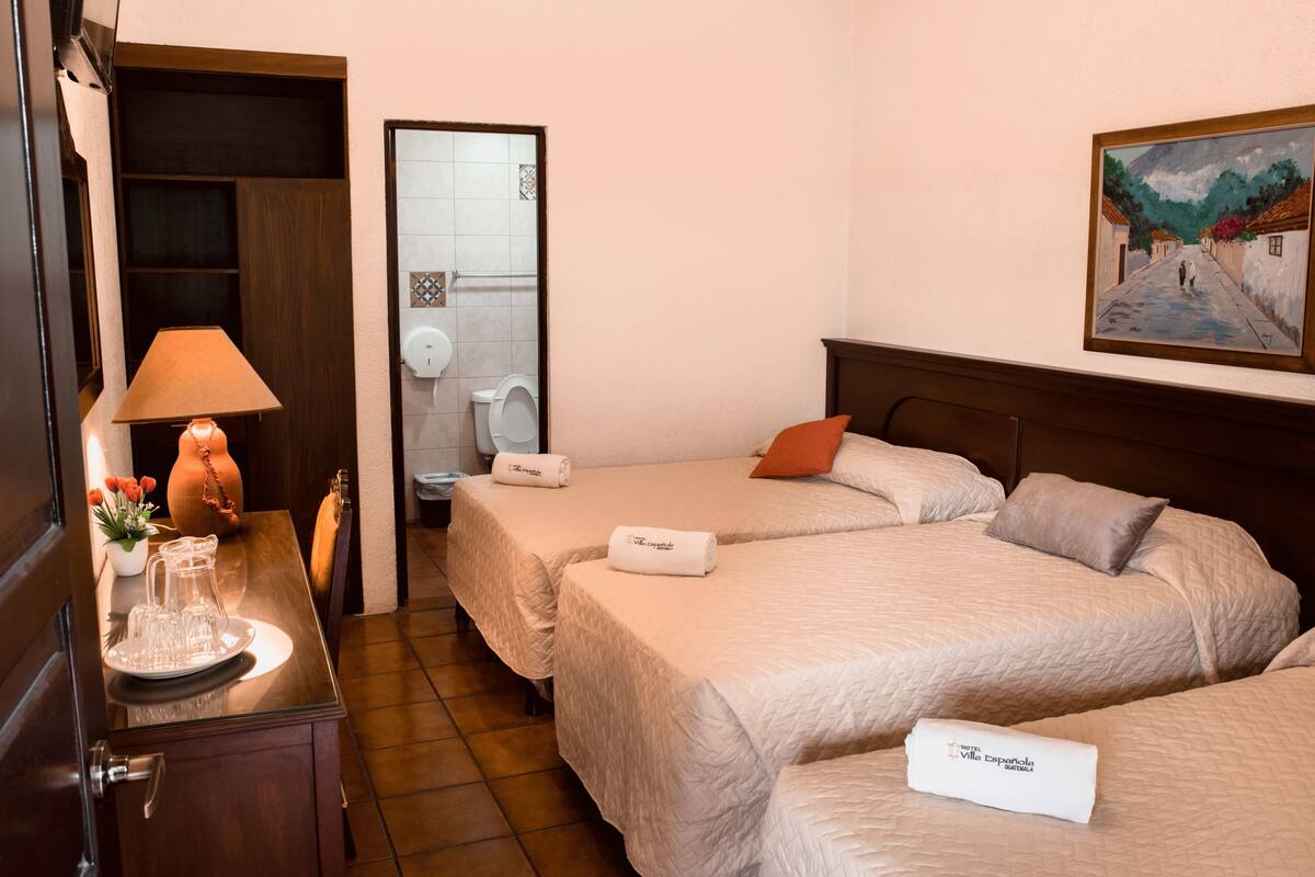 A hotel room with two beds, a wooden desk with a lamp, a vase with flowers, and a painting on the wall.
