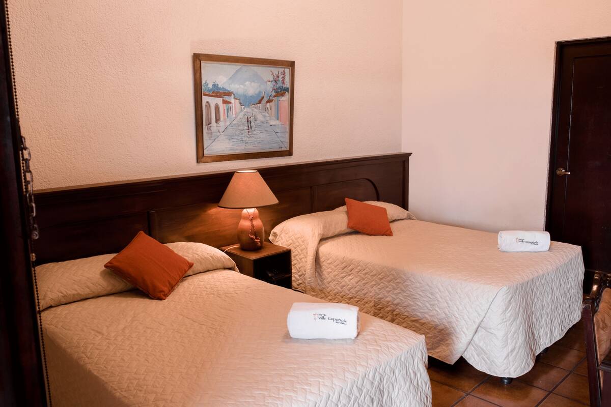 A hotel room with two beds, a nightstand with a lamp, a painting on the wall, and a door.
