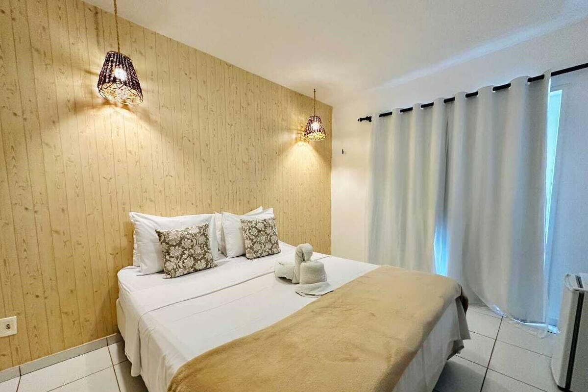 Une chambre d’hôtel avec un mur en bois, un lit avec une literie blanche, deux lampes fixées au mur et une fenêtre avec des rideaux.