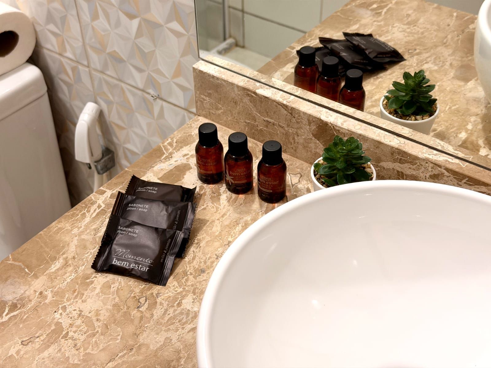 Une salle de bain avec un comptoir en marbre, un lavabo blanc, une petite plante en pot et plusieurs articles de toilette, notamment des savons et des bouteilles de shampoing.