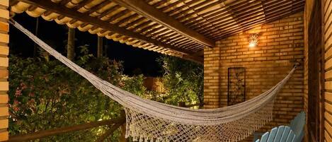 A hammock under a wooden pergola, two light blue chairs, and a brick wall with a lit lantern.