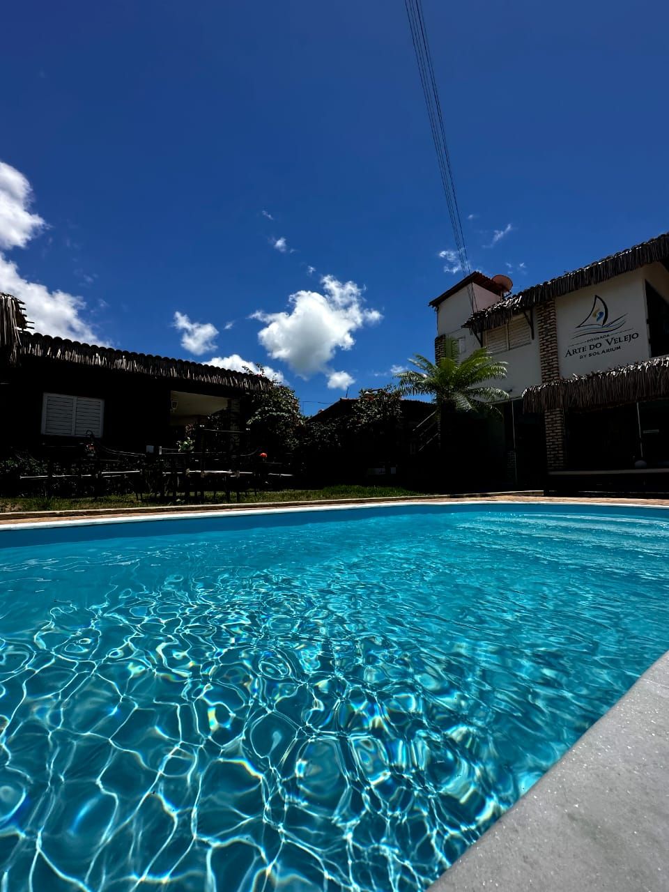 A swimming pool with clear water, surrounded by buildings with thatched roofs and a clear blue sky.
