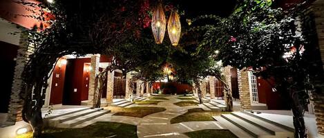 Un camino conduce a un edificio con varias puertas, rodeado de árboles e iluminado por luces superiores.