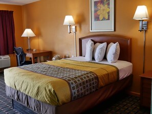 A hotel room with a large bed, two bedside lamps, a desk with a chair, a telephone, and a framed picture on the wall.