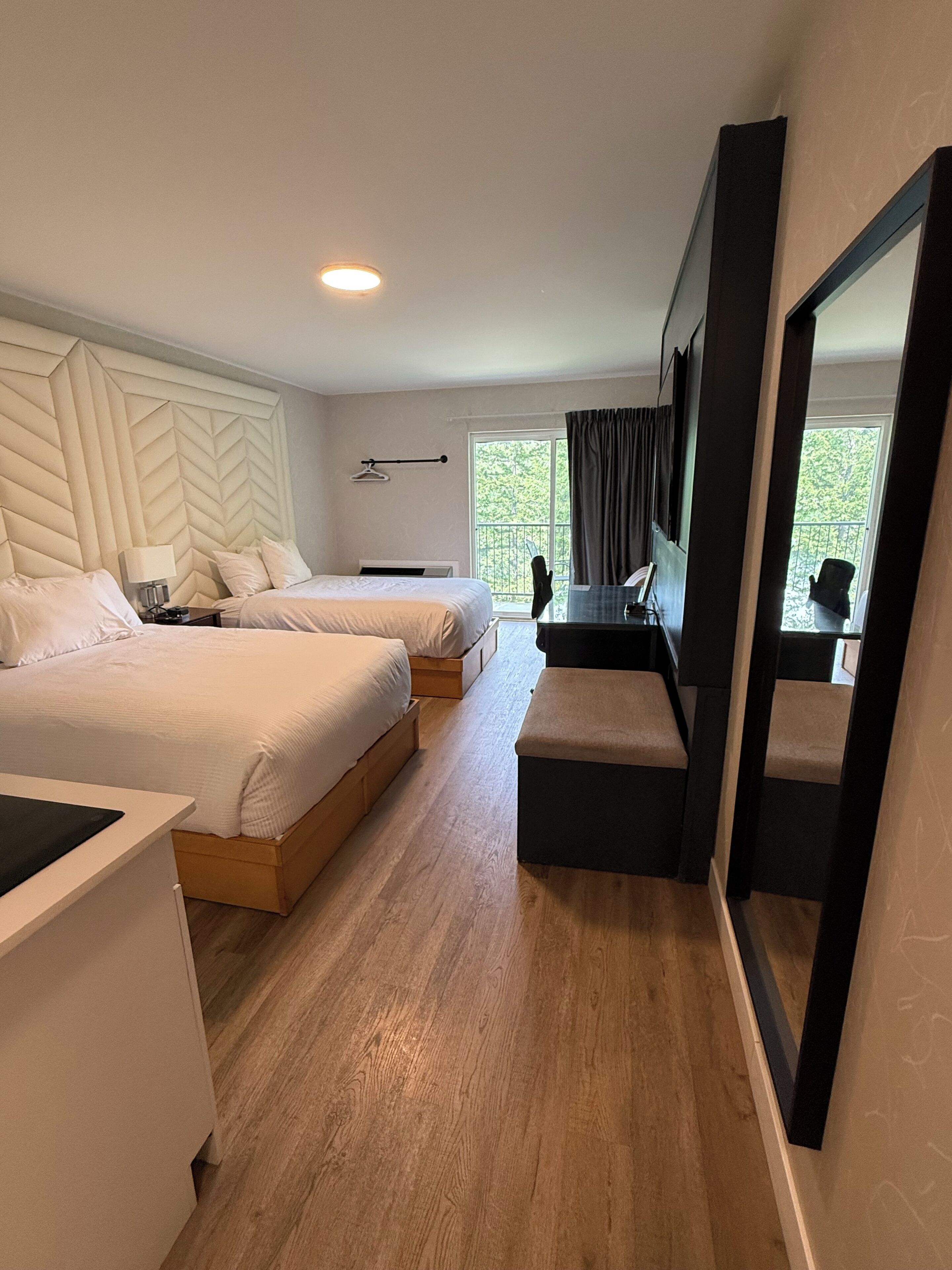 A hotel room with two beds, a desk, a chair, a mirror, and a window with a view of greenery.