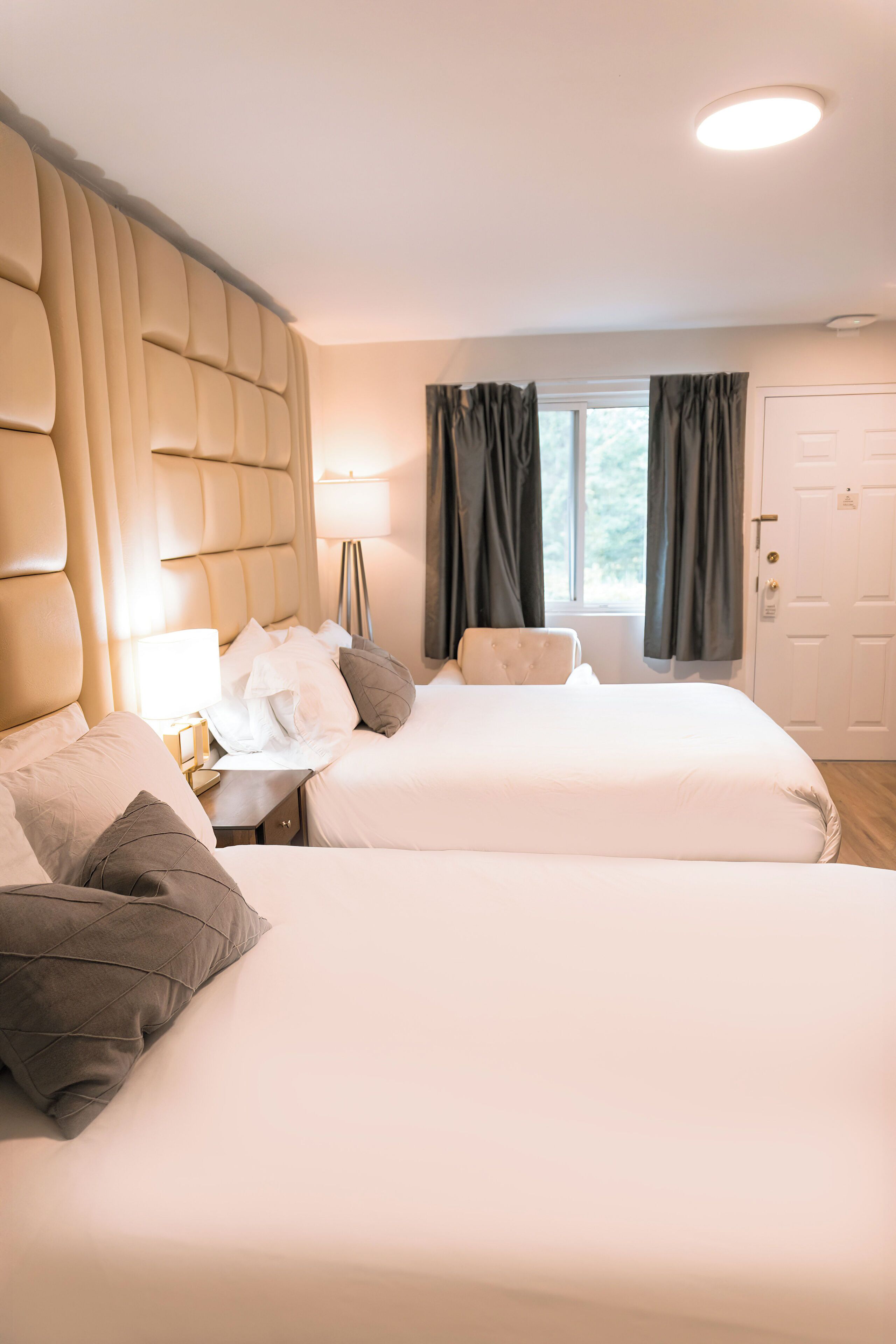 Une chambre d’hôtel avec deux lits, une tête de lit, une table de chevet avec une lampe et une fenêtre avec des rideaux.