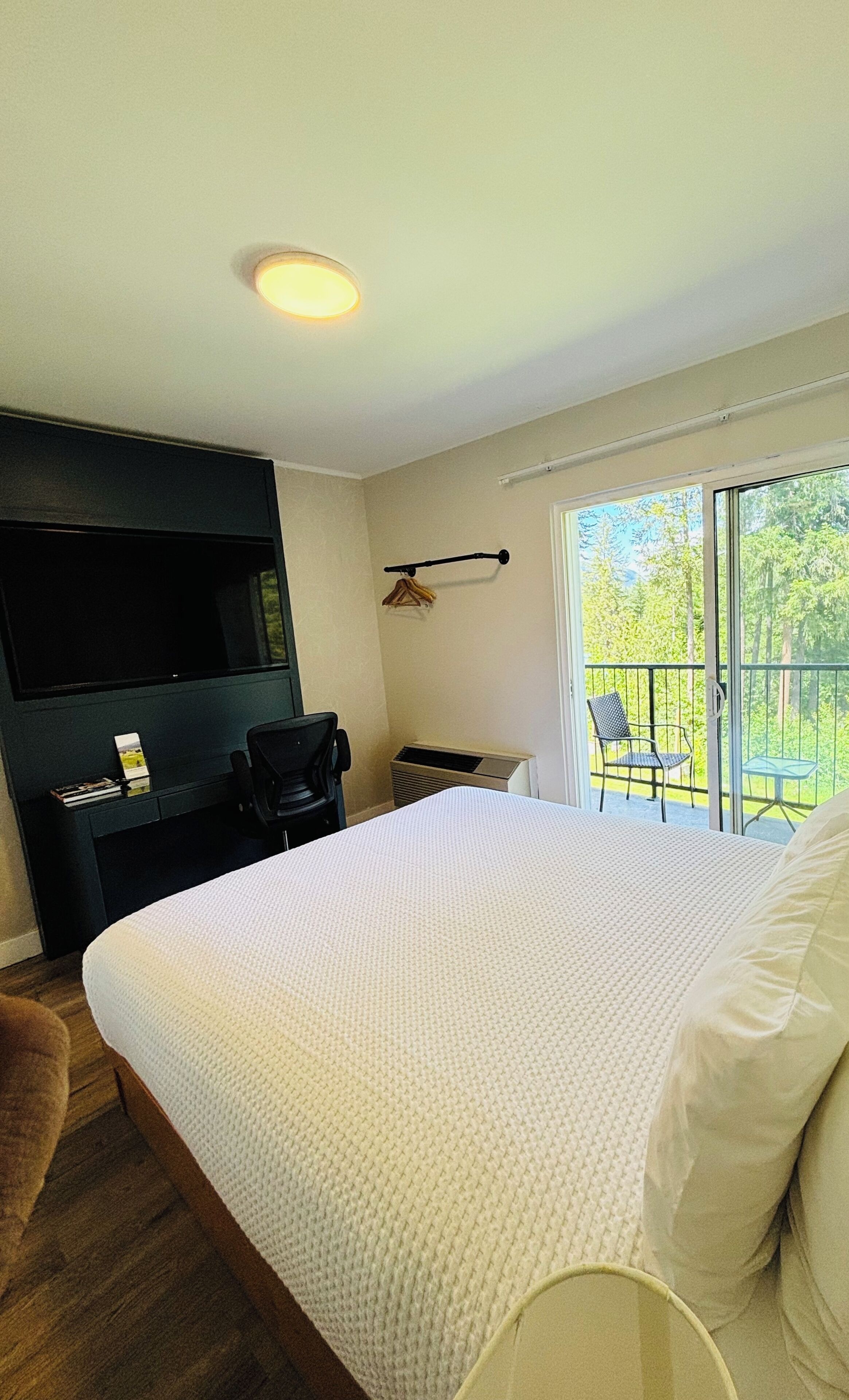 A bedroom with a bed, a TV, a chair, a small table, and a balcony with a view of trees.