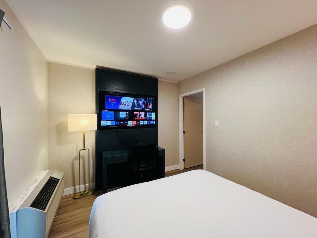 Ein Hotelzimmer mit einem Bett, einem Schreibtisch mit Computer, einem Fernseher, der mehrere Kanäle zeigt, und einer Lampe.