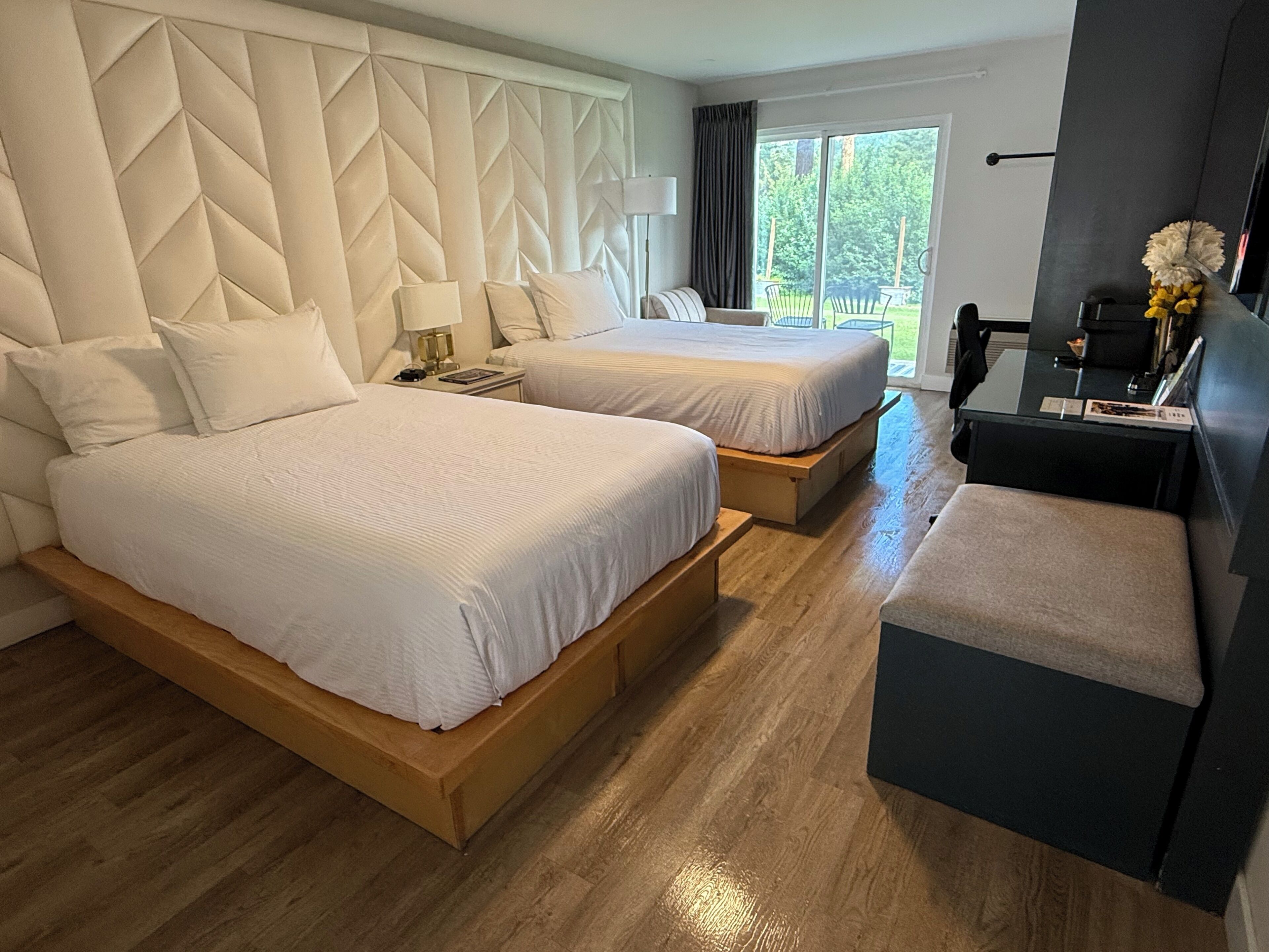 A hotel room with two beds, a desk, a chair, and a view of the outdoors.